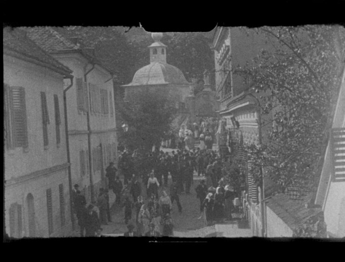 A black and white photo shows people on a narrow street in the city with a church behind them. The people are leaving the church.