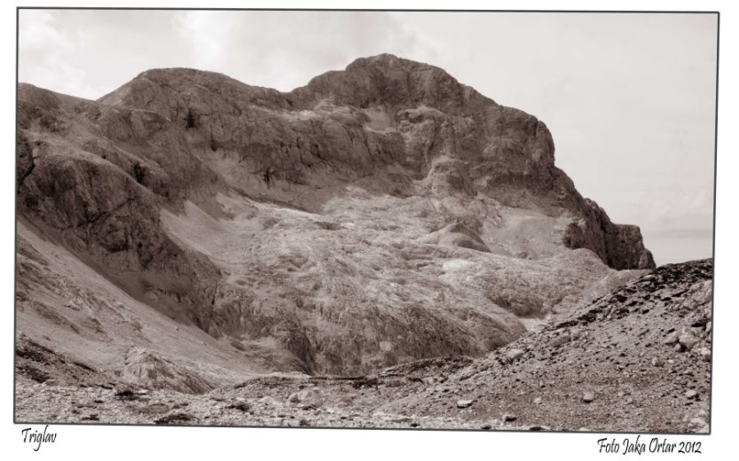 A photo made in retro style depicts steep, rocky mountain slope beneath Triglav, Slovenia’s highest peak, with a patch of snow and ice.
