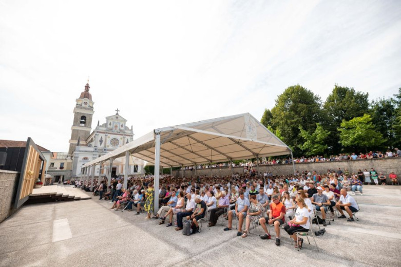 The photo shows a large crowd of people seated under a big white outdoor tent, attending an event in front of a church with a tall bell tower. More people are standing along the wall surrounding the venue, and it is a sunny day.