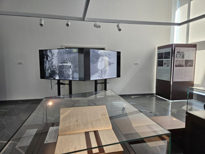 Museum exhibition room with glass display cases containing old documents. A black-and-white film scene is the two TV screens in the background. 