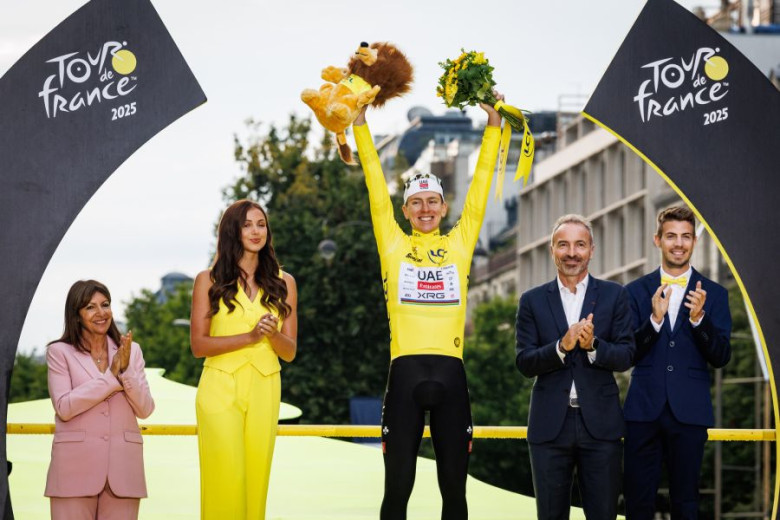 A photo of Tadej Pogačar on the top Tour de France podium in a yellow jersey, holding a stuffed lion toy in his right hand and a bouquet in his left, with two men and two women clapping beside him.