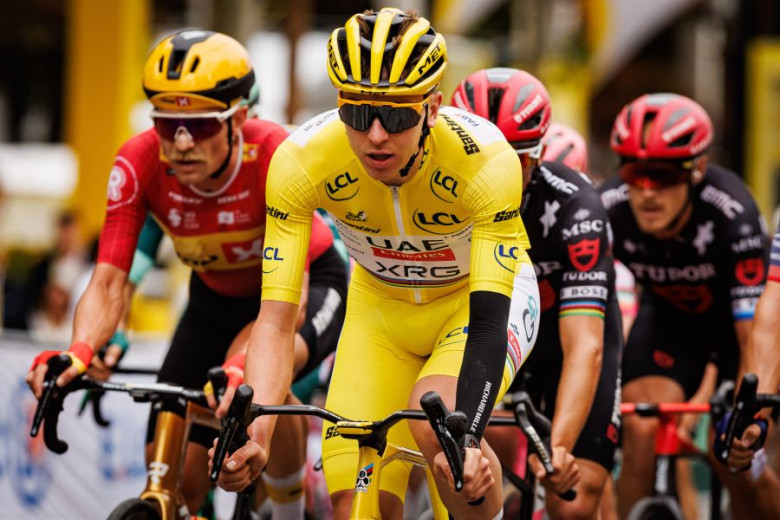 A group of cyclists in full racing gear — helmets, glasses, and jerseys — led by Tadej Pogačar in the yellow jersey at the front.