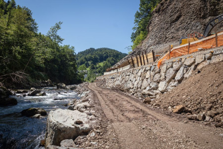 The image shows a partially reconstructed dirt road running alongside a fast-flowing stream in a forested, mountainous area. On the right, a stone-reinforced slope and construction machinery are visible, indicating ongoing repair work.
