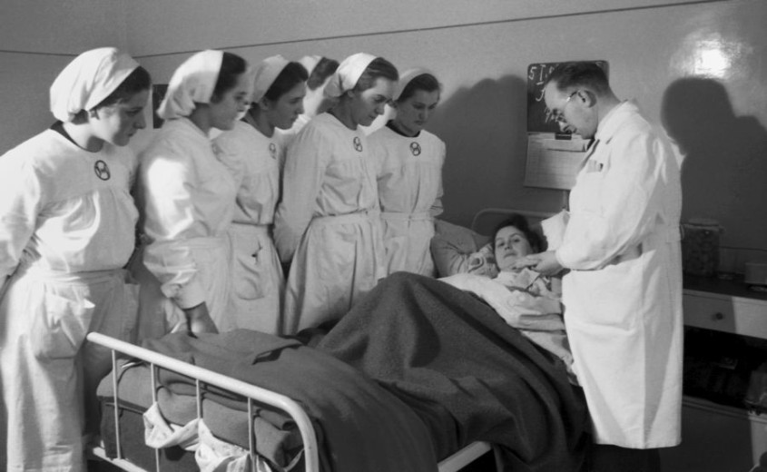 Skupina medicinskih sester in zdravnik stoji ob bolniški postelji ter opazuje pacientko, ki leži pod odejami. Zdravnik očitno govori ali nekaj razlaga medicinskim sestram.