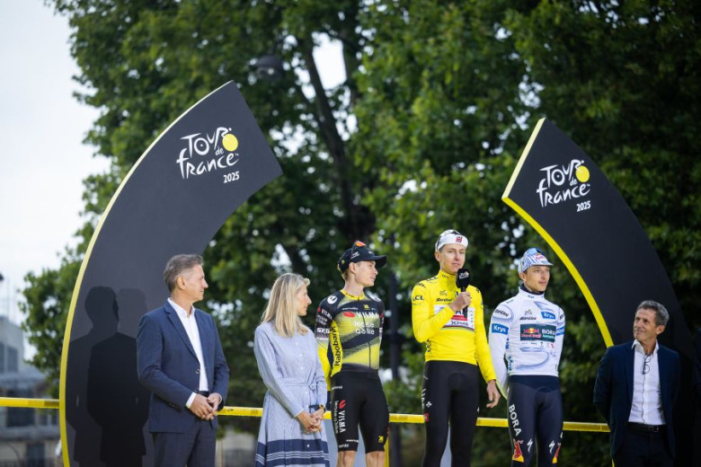 The photo shows the top three cyclists of the 2025 Tour de France standing on a podium outdoors, with Tadej Pogačar in the center wearing the yellow jersey and speaking into a microphone. 