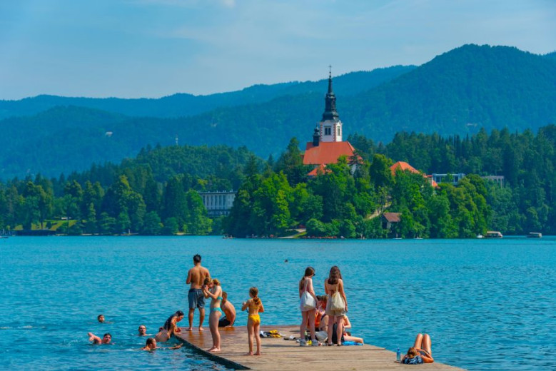 Fotografija prikazuje živahno dogajanje ob obali Blejskega jezera, kjer ljudje počivajo na pomolu in plavajo v kristalno čisti vodi, v ozadju pa se na slikovitem otoku vidi cerkev.