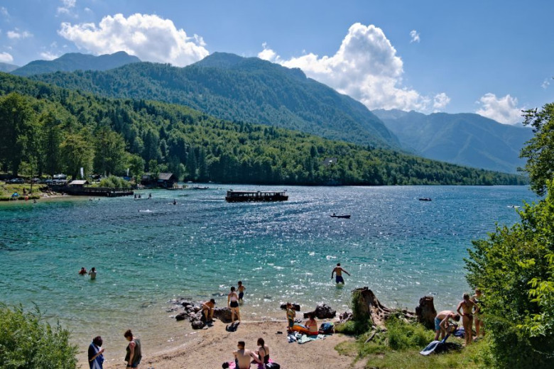 Fotografija prikazuje pogled na obalo Bohinjskega jezera, kjer ljudje uživajo na plaži in se kopajo, v ozadju pa se razprostira gozd in mogočne gore.