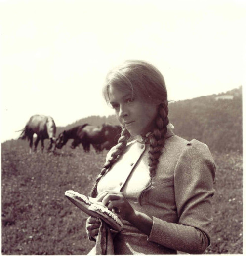 An old black-and-white photo of a woman holding a honeybread heart. She has her hair braided into two plaits and stands in a meadow, with two cows visible in the background.