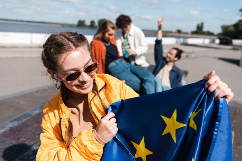 A young woman with dark hair and sunglasses holding the EU flag in her hands. In the background we can see more young people.