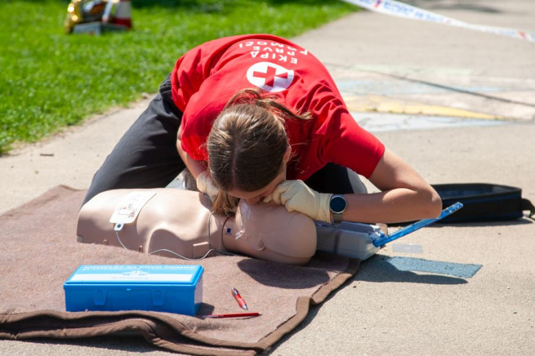 Red Cross volunteer practices CPR rescue breaths on a mannequin outdoors with first aid kit during emergency training.