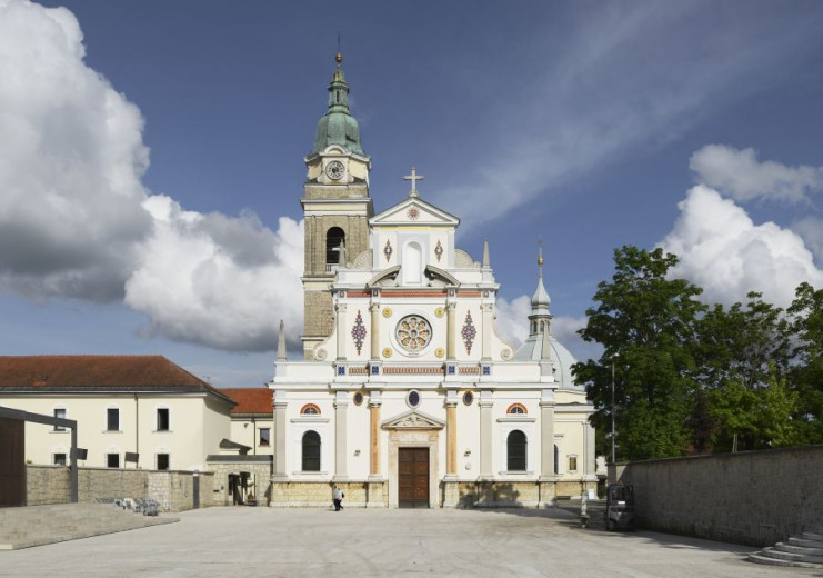 This photo shows the front view of a large church with an ornate white façade, decorative details, and a tall bell tower topped with a green dome. In front of the church is a wide open square under a bright sky with scattered clouds.