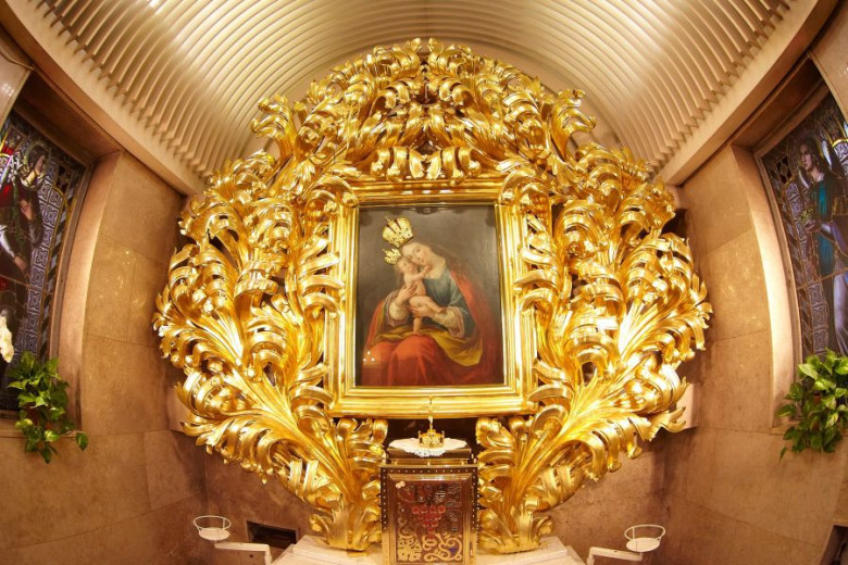 This photo shows a richly decorated golden altar with an ornate frame surrounding a painting of the Virgin Mary holding the infant Jesus. The scene is flanked by stained-glass windows on both sides, adding to the sacred atmosphere.