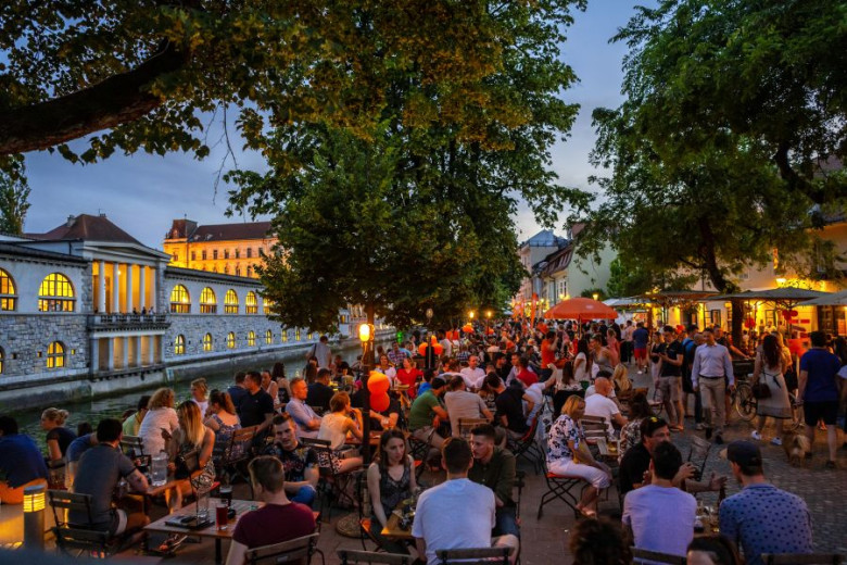 This photo shows crowds of people gather at outdoor cafes and restaurants along a riverside promenade in the evening, under trees and warm lights, with historic buildings illuminated in the background.