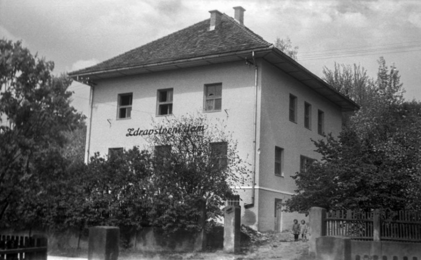 Črno-bela fotografija dvonadstropne stavbe z napisom "Zdravstveni dom", obdane z drevesi in ograjo; pri vhodnih vratih stojita dva otroka.