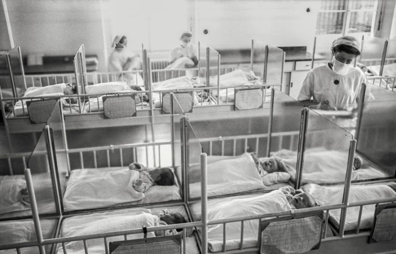 Črno-bela fotografija novorojenčkov v majhnih posteljicah, postavljenih drug ob drugem, med katerimi tri medicinske sestre nadzorujejo in držijo nekatere novorojenčke.
