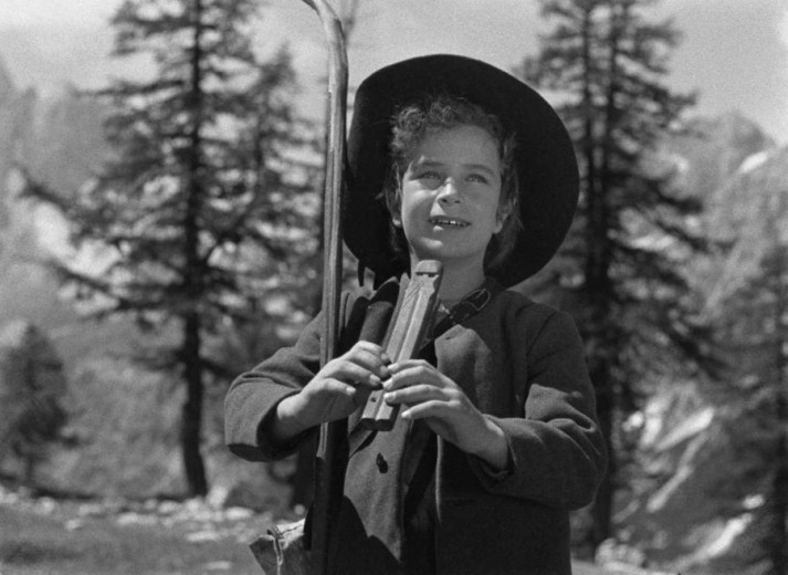 This black-and-white photo shows a young boy standing in front of a backdrop of mountains and trees. He is dressed in a traditional Alpine uniform, wearing a hat and holding a flute in his hand