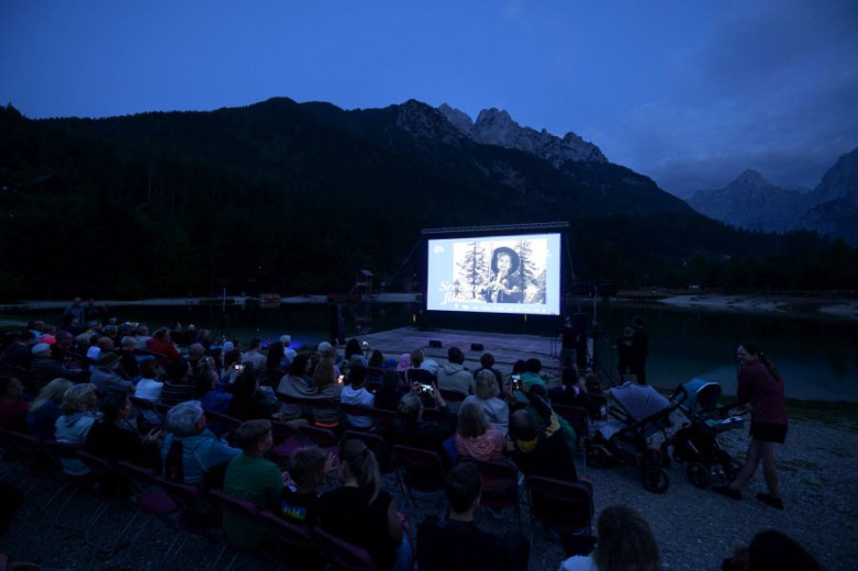 A nighttime photo of an open-air cinema shows a movie screen in the center with mountains rising in the background. In the darkness, rows of viewers can be seen watching the film.