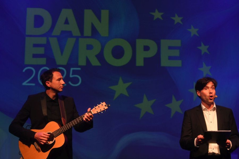 A man holding a guitar and another man talking on the microphone on a stage. In the backgroung is a title Europe day with a circle of twelve golden stars on a blue background.