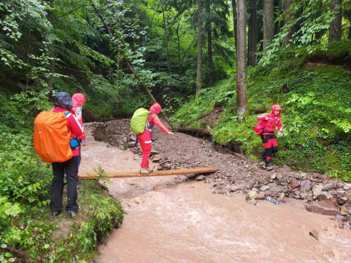 This photo shows four rescuers carefully crossing a muddy stream on a narrow wooden board, each carrying a large backpack.