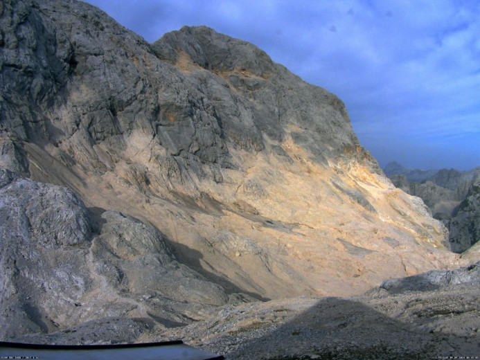 The photo shows a steep, rocky mountain slope beneath Triglav, Slovenia’s highest peak, with little, barely visible patches of ice. The sky is partialy cloudy.