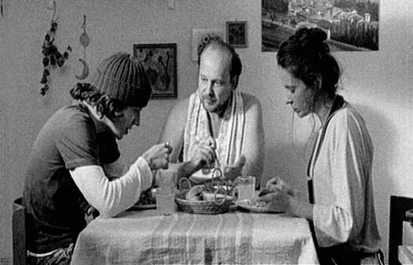A black-and-white photo of three people sitting around a kitchen table, sharing lunch. In the center is a man with only a towel draped around his neck. To his left sits a younger man wearing a woolen hat, and to his right, a woman with dark hair.