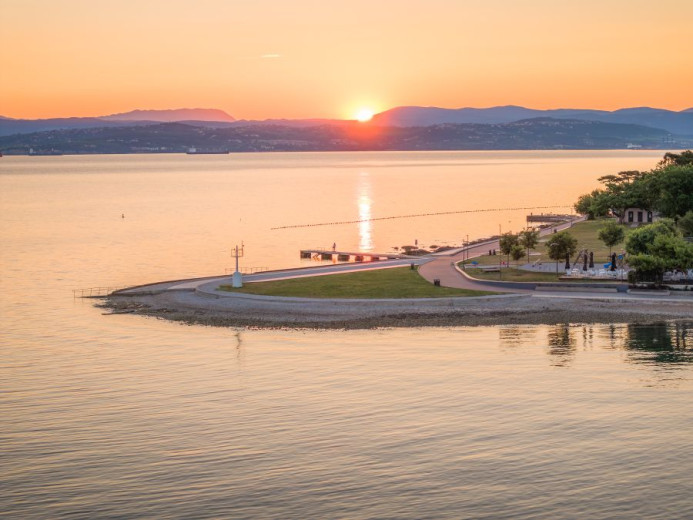 Fotografija sončnega zahoda z gorami na obzorju, v ospredju pa morje in rt s plažo. 