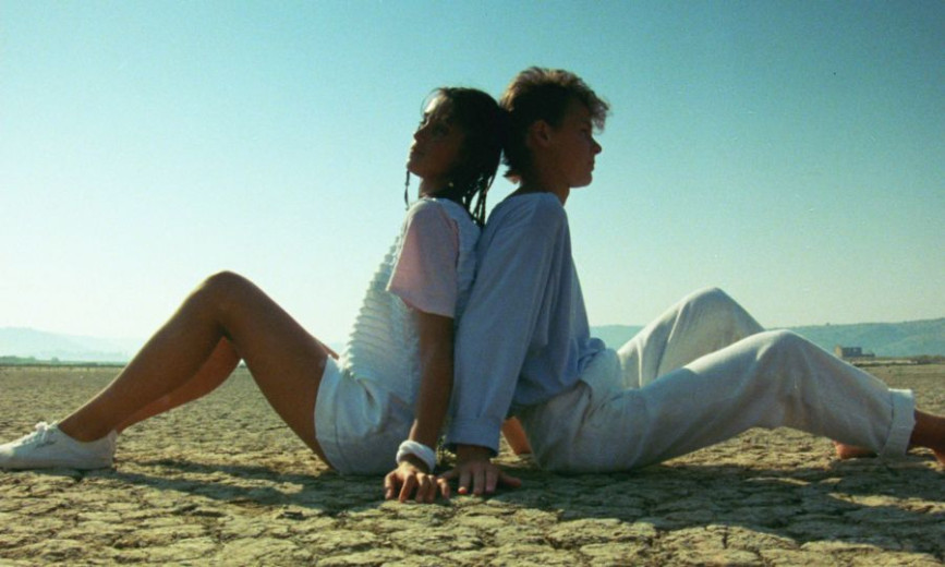 A photo of a teenage boy and girl sitting back-to-back on the dirt ground, with nothing but clear blue skies above them.