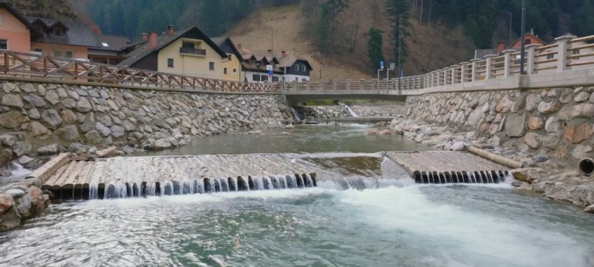 This photo shows a river channel renovated after the flood, with reinforced banks made of stone and concrete. Clear water now flows through it, with houses lining the edge of the river.