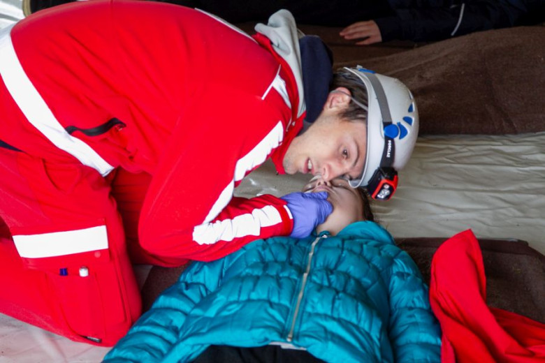 A first responder, wearing a red uniform and headlamp, examines an unresponsive patient lying on a blanket. 