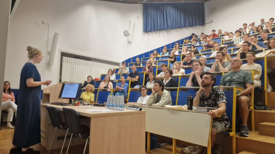 A photo woman stands and speaks at the front of a lecture hall, addressing an audience of seated adults who are attentively listening. A presentation is displayed on a computer screen beside her.