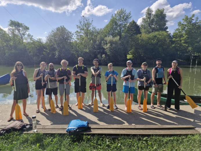 This photo shows a group of teenagers standing on the bank of a river, holding holding a paddle in their hands. There is a canoe in the water in the background.