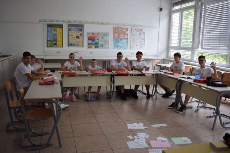 This photo shows a classroom with teenage students sitting behind desks full of papers and notebooks, waving at the camera. There are also some papers on the floor.