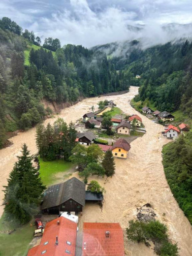 This aerial photo shows a flooded village nestled in a steep, forested valley. A muddy, fast-moving river has overflowed, submerging roads and reaching the walls of many houses.