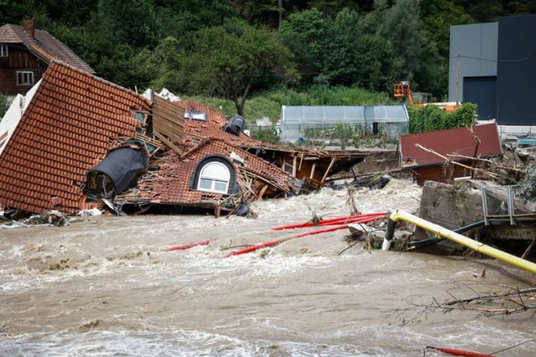 This picture shows a house that is flooded and damaged. The muddy water is so high that only the roof is visible, and it appears to have been partially washed away by the raging current.