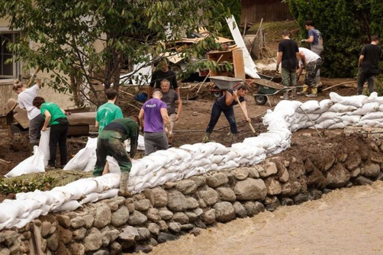 This photo shows people on a river bank filling sanbags and piling them up just next to the river. They are wearing rubber boots and work clothes and some are covered in mud. 