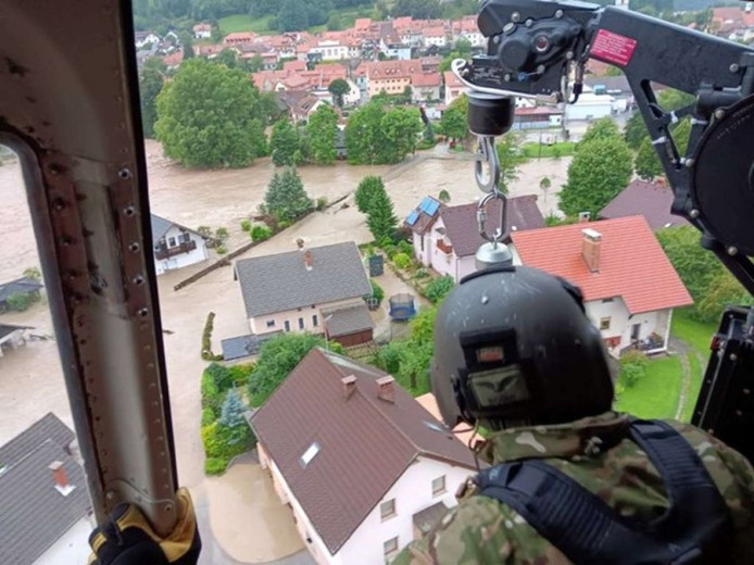 An aerial photograph taken from a Slovenian Armed Forces helicopter shows a village completely surrounded by muddy floodwaters, with houses isolated like small islands.