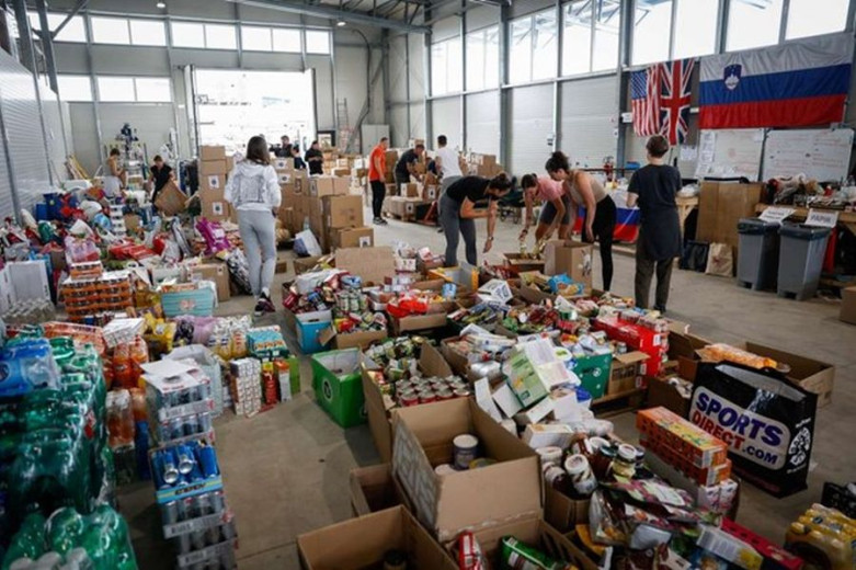 This photo shows a big storage room, full of boxes of water and canned drinks, different types of packed food and volunteers preparing the food packages.