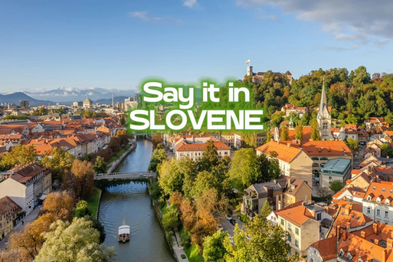 An aerial photo of Ljubljana on a sunny day, with Ljubljanica river, the old town and castle in the backround. In the middle of the photo there is a sign Say it in SLOVENE.