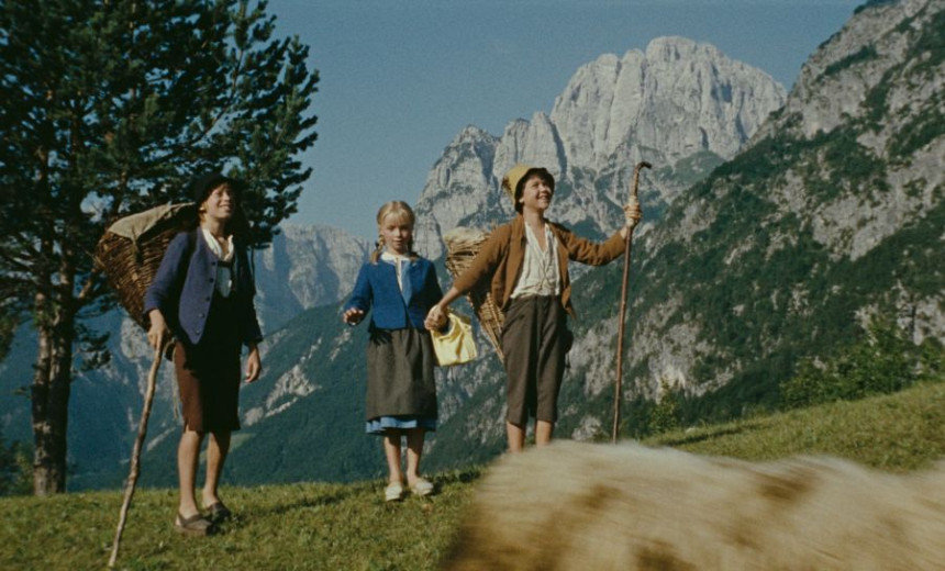 This photo shows a young girl standing in the mountains, with two young boys by her side. The boys are holding shepherd’s staffs, wearing traditional Alpine hats, and carrying woven baskets on their backs. The girl is wearing a blue dress.