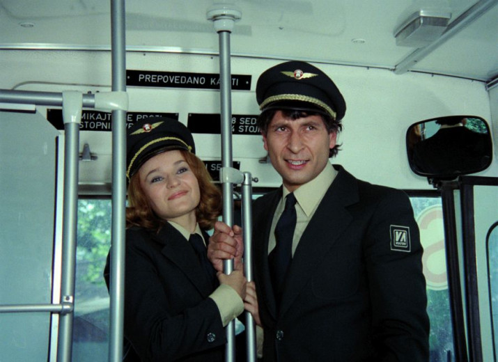 A photo of a man and a younger woman dressed in dark blue bus driver uniforms with hats, standing on a bus.