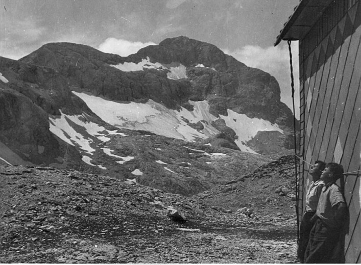 Two young boys standing at the side of a mountain hut (Stanič Hut) looking in the distance. With mountain slopes partially covered with snow in the background.