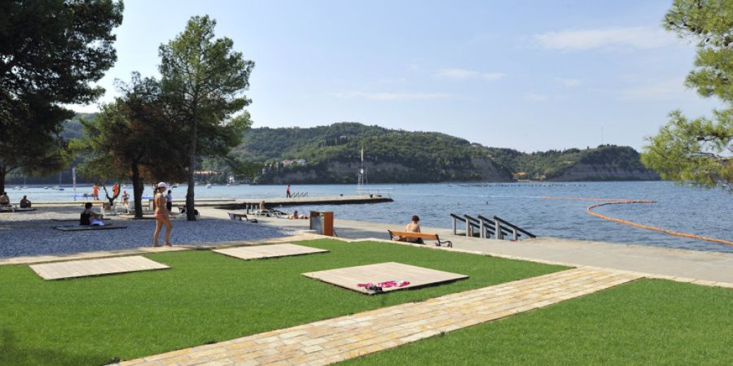 Fotografija plaže, ki je delno prekrita z travo in delno prodnata. Na plaži so ljudje v kopalkah. S plaže je razgled na pečine na drugi strani zaliva.