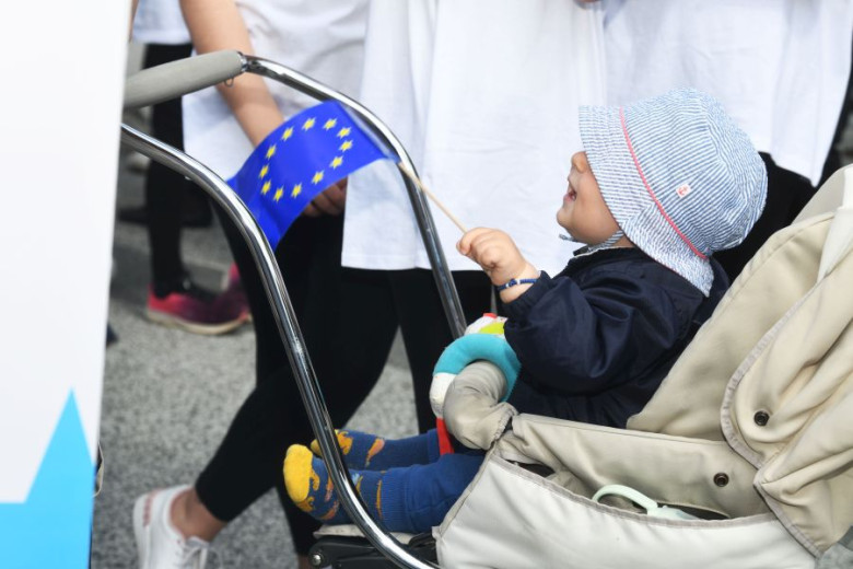 A baby in a stroller holding a flag featuring a circle of twelve golden stars on a blue background.