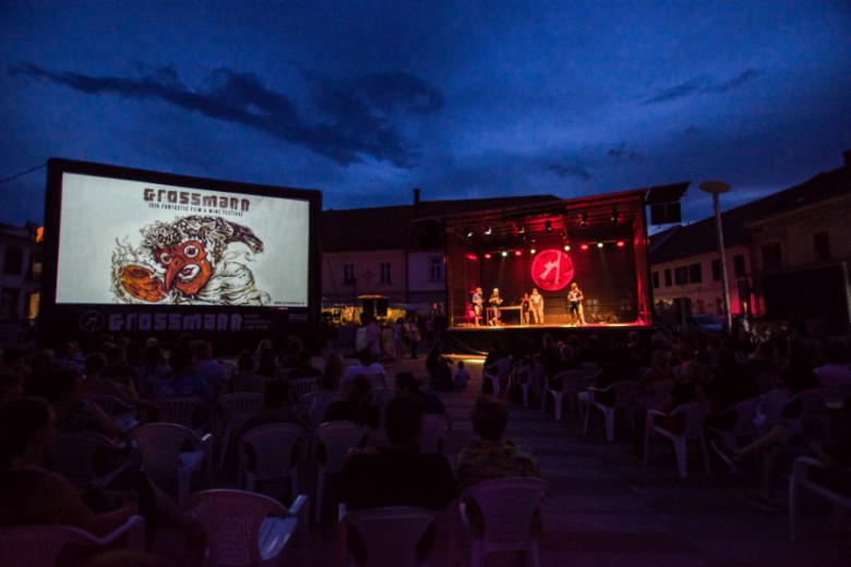 This photo captures an open-air movie screening at night. On the left, a large screen displays an animated image of a man wearing a red mask with a long nose, with the word "Grossmann" above him. On the right, a stage is set up with five people standing. 