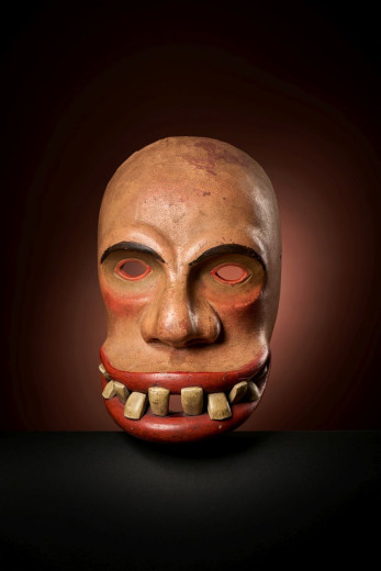 A bald carnival mask with hollow eyes, red lips, and uneven teeth on a dark background.