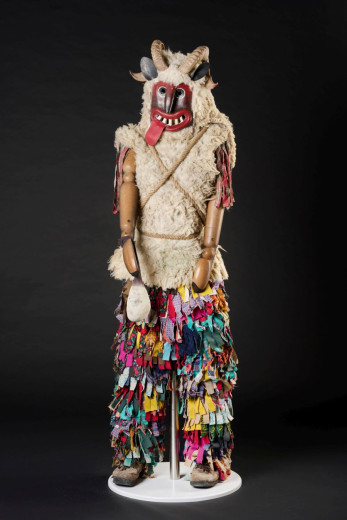 A full-body carnival costume with a horned mask, red mouth, fur vest, and colorful fabric strips.