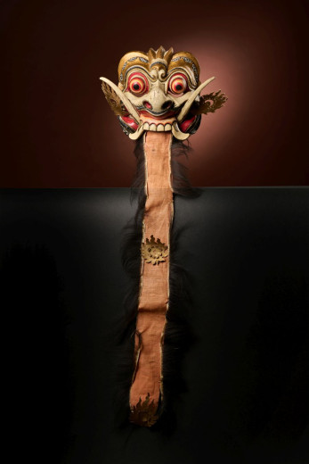 A carved carnival mask with wide eyes and fangs, mounted on a long wooden handle.