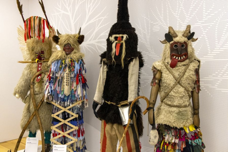 Four carnival costumes with horned masks, fur garments, bells, and colorful fabric strips stand side by side.