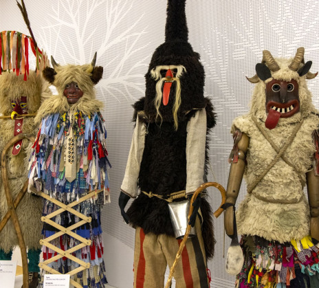 Four carnival costumes with horned masks, fur garments, bells, and colorful fabric strips stand side by side.