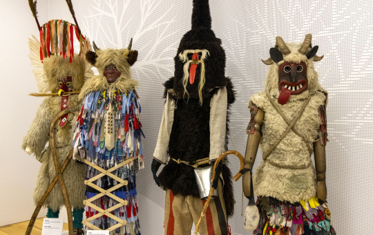 Four carnival costumes with horned masks, fur garments, bells, and colorful fabric strips stand side by side.